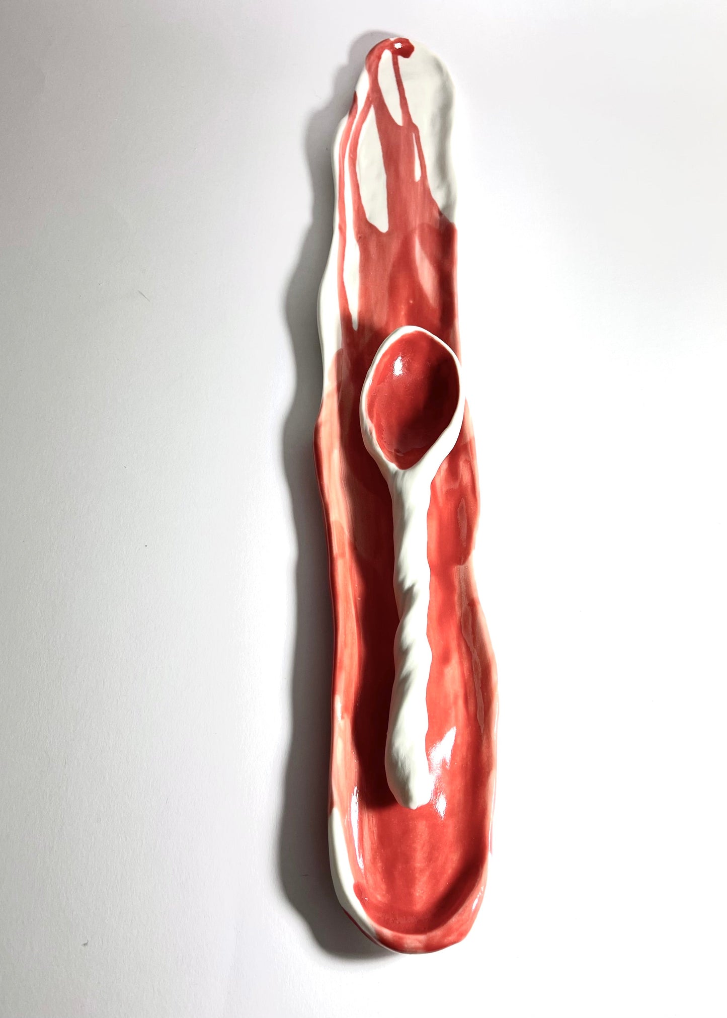 A sculptural ceramic dish featuring a glossy red and white abstract design with a built-in spoon, photographed against a plain white background.
