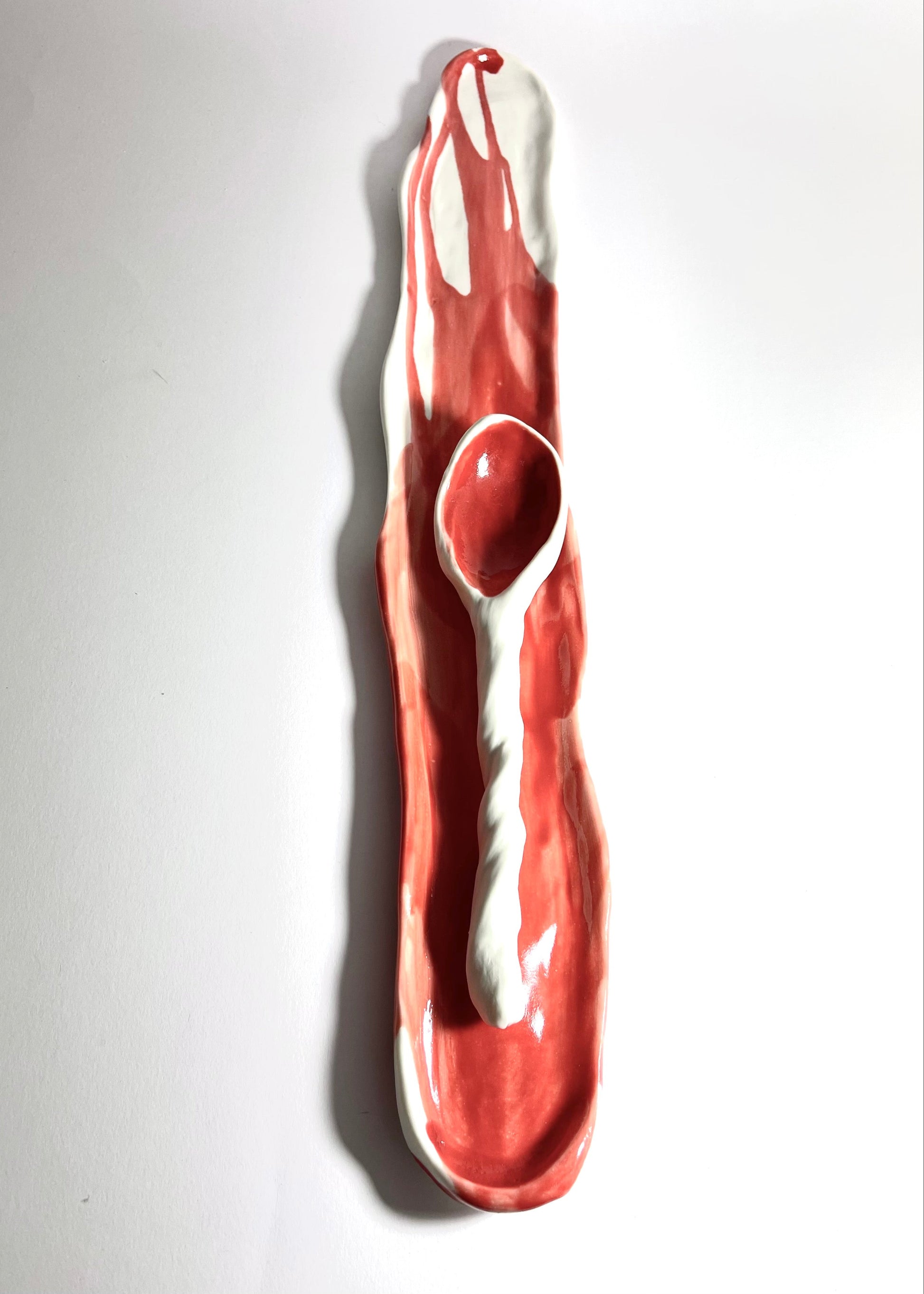 A sculptural ceramic dish featuring a glossy red and white abstract design with a built-in spoon, photographed against a plain white background.