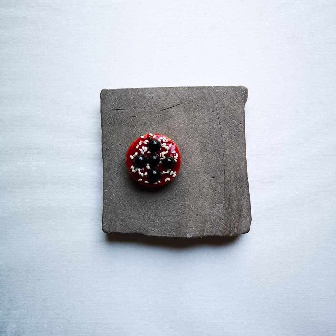 A small round red button with white and black speckles, placed on a square textured gray slate coaster against a light gray background.