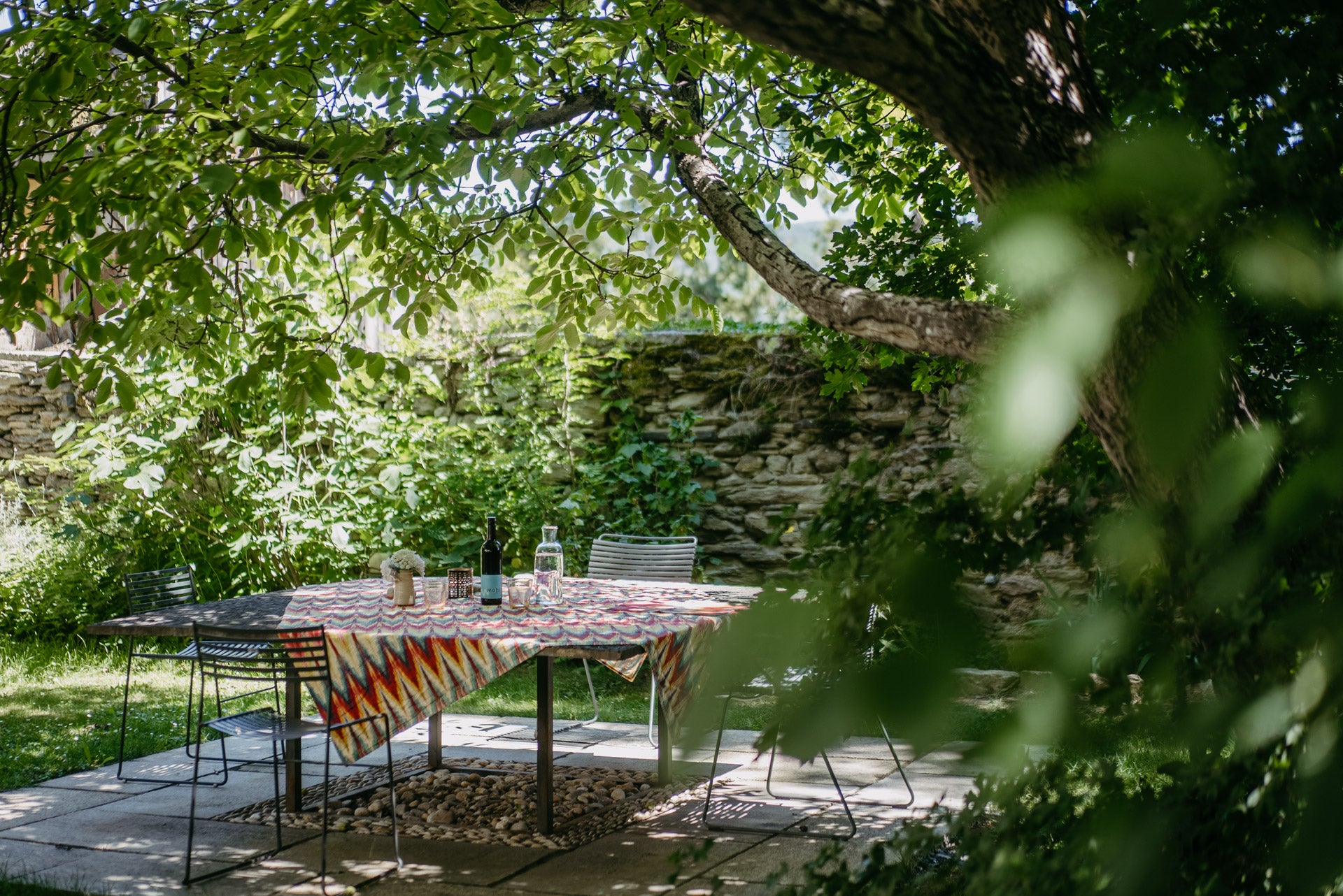 A rectangular outdoor dining table with a colorful geometric patterned tablecloth, set on a stone patio under a leafy tree with a stone wall in the background.