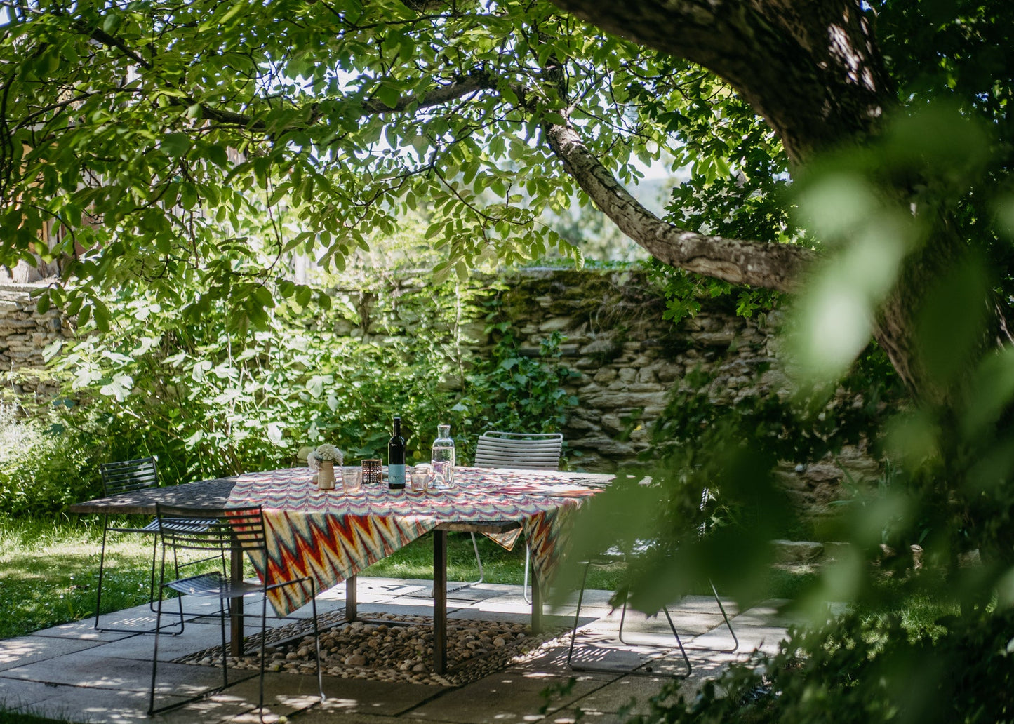 A rectangular outdoor dining table with a colorful geometric patterned tablecloth, set on a stone patio under a leafy tree with a stone wall in the background.