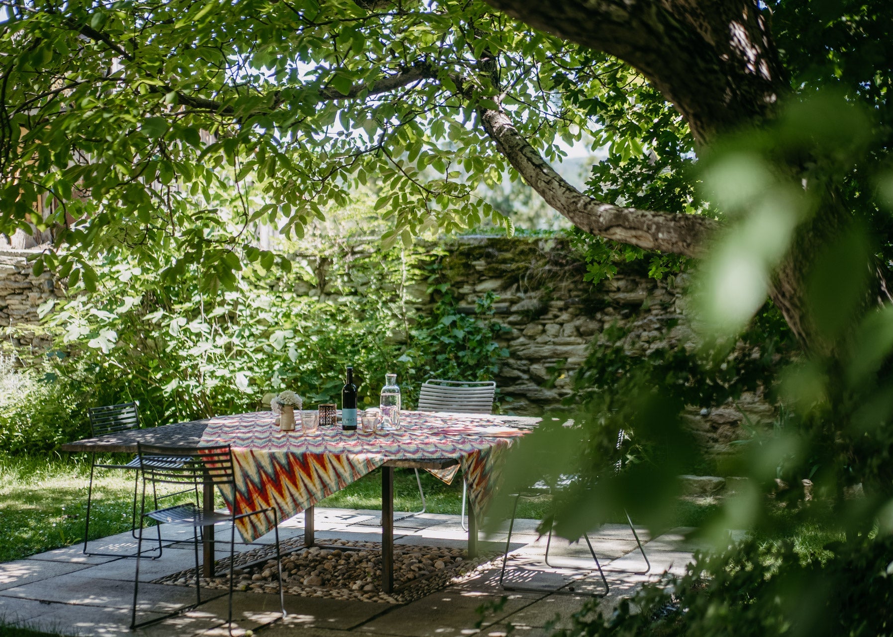 A rectangular outdoor dining table with a colorful geometric patterned tablecloth, set on a stone patio under a leafy tree with a stone wall in the background.
