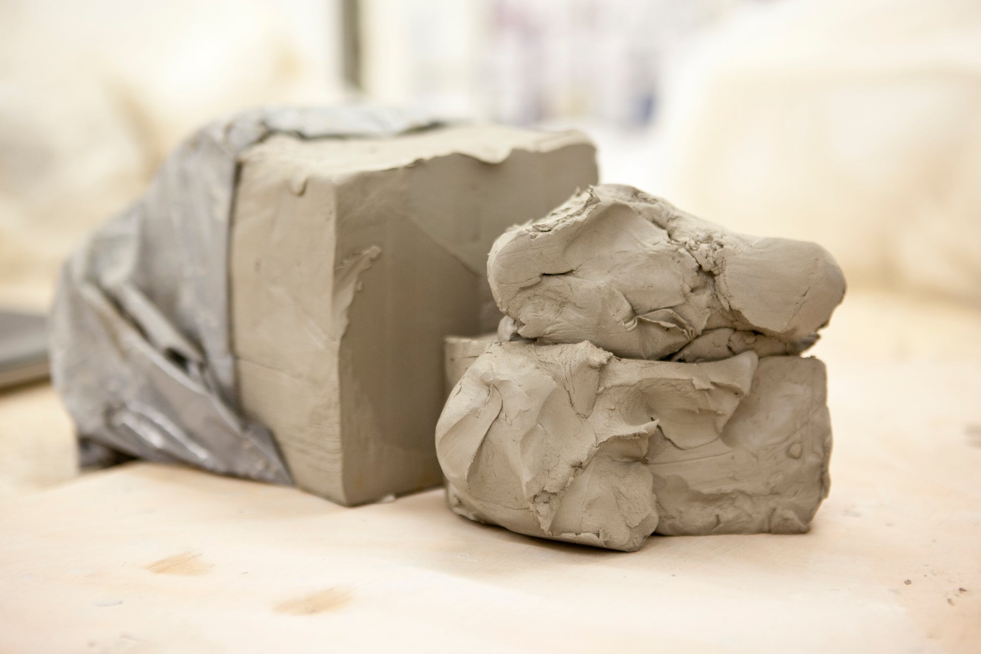 Two mounds of gray clay, one wrapped in a translucent plastic sheet and one unwrapped, resting on a light beige surface with a softly blurred background.