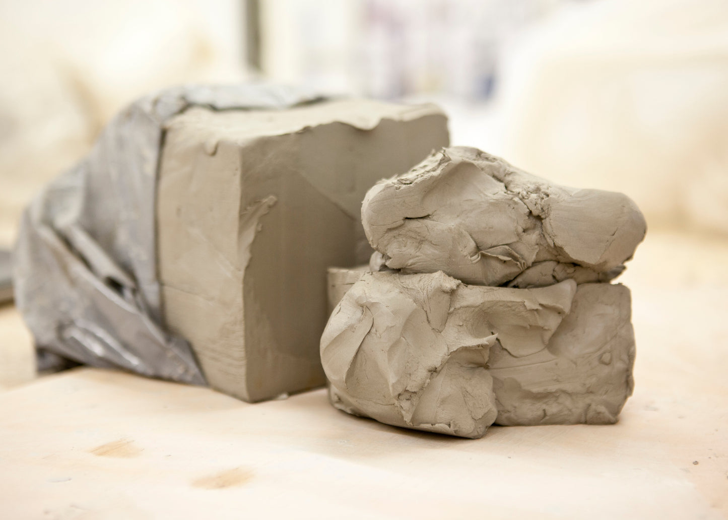 Two mounds of gray clay, one wrapped in a translucent plastic sheet and one unwrapped, resting on a light beige surface with a softly blurred background.