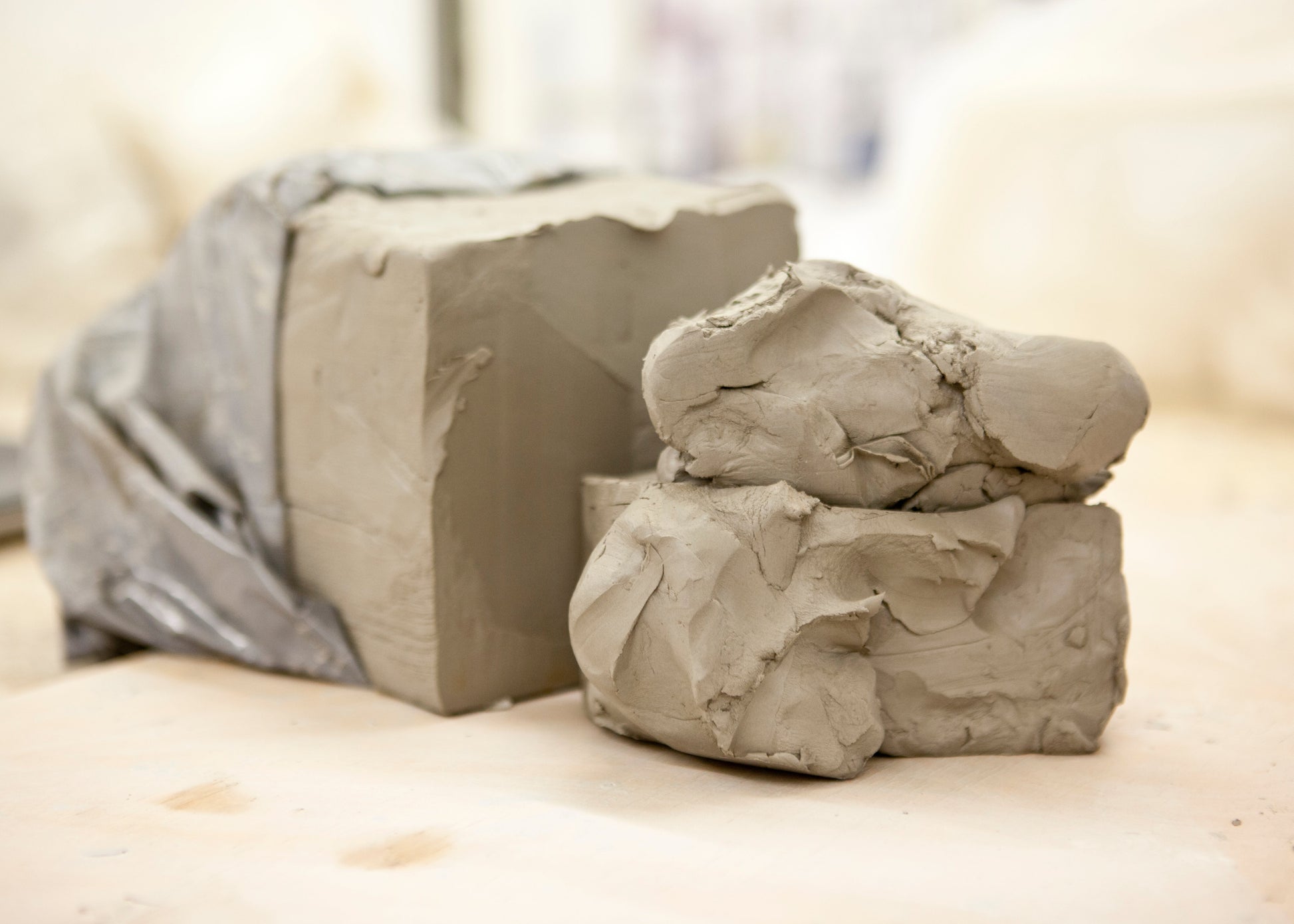 Two mounds of gray clay, one wrapped in a translucent plastic sheet and one unwrapped, resting on a light beige surface with a softly blurred background.