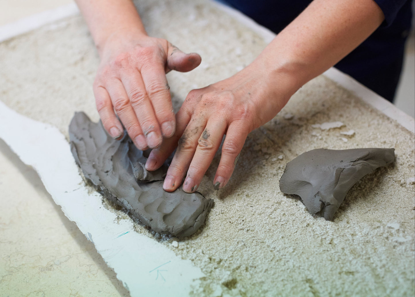 Close-up of a person's hands shaping a piece of gray clay on a textured beige surface.