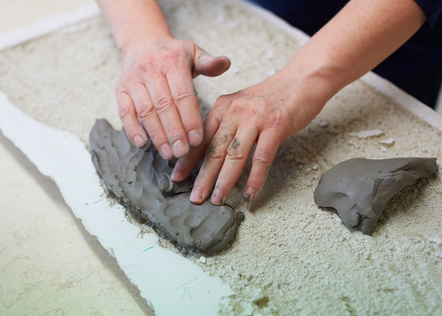 Close-up of a person's hands shaping a piece of gray clay on a textured beige surface.