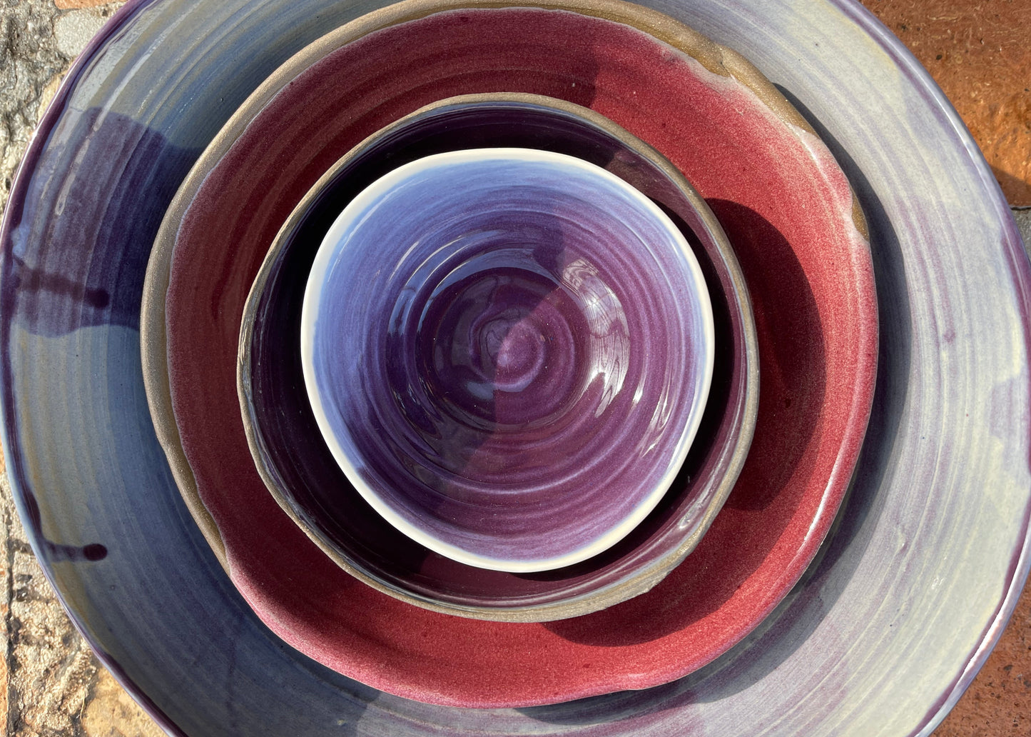 A set of nested ceramic bowls and plates with a hand-painted glaze featuring shades of purple, burgundy red, and slate blue, photographed from directly above.