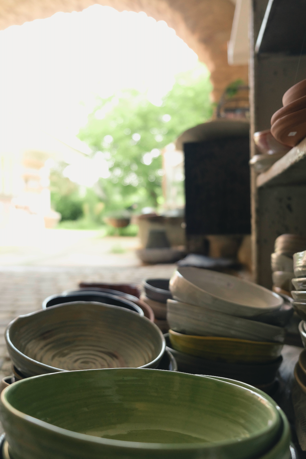 A close-up, shallow depth-of-field view of handmade ceramic bowls and plates in various earthy tones, with a prominent green bowl in the foreground and a soft-focus outdoor patio scene in the background.