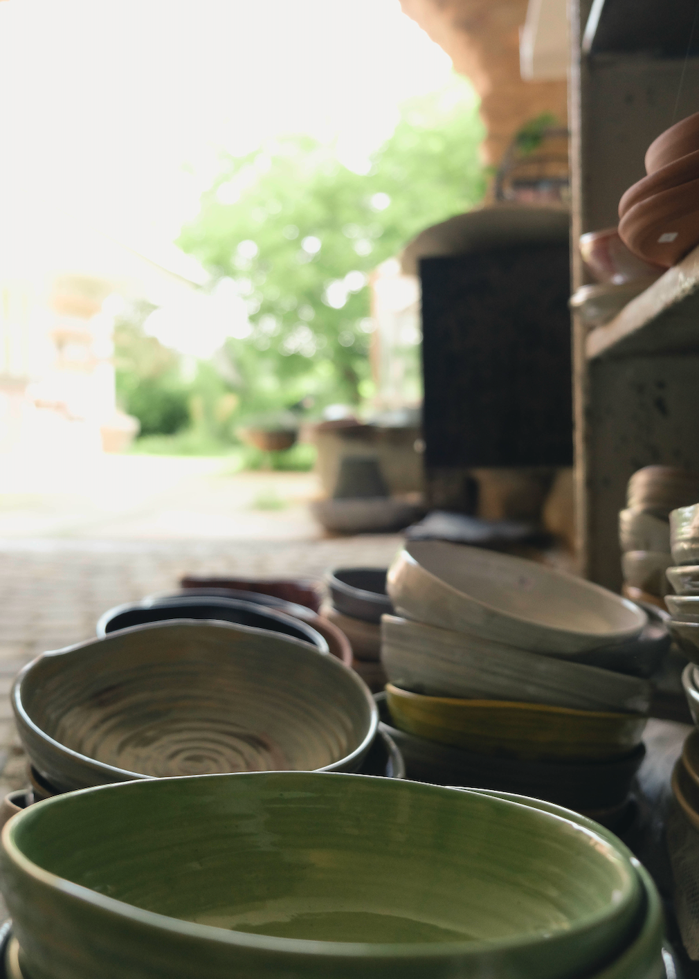 A close-up, shallow depth-of-field view of handmade ceramic bowls and plates in various earthy tones, with a prominent green bowl in the foreground and a soft-focus outdoor patio scene in the background.