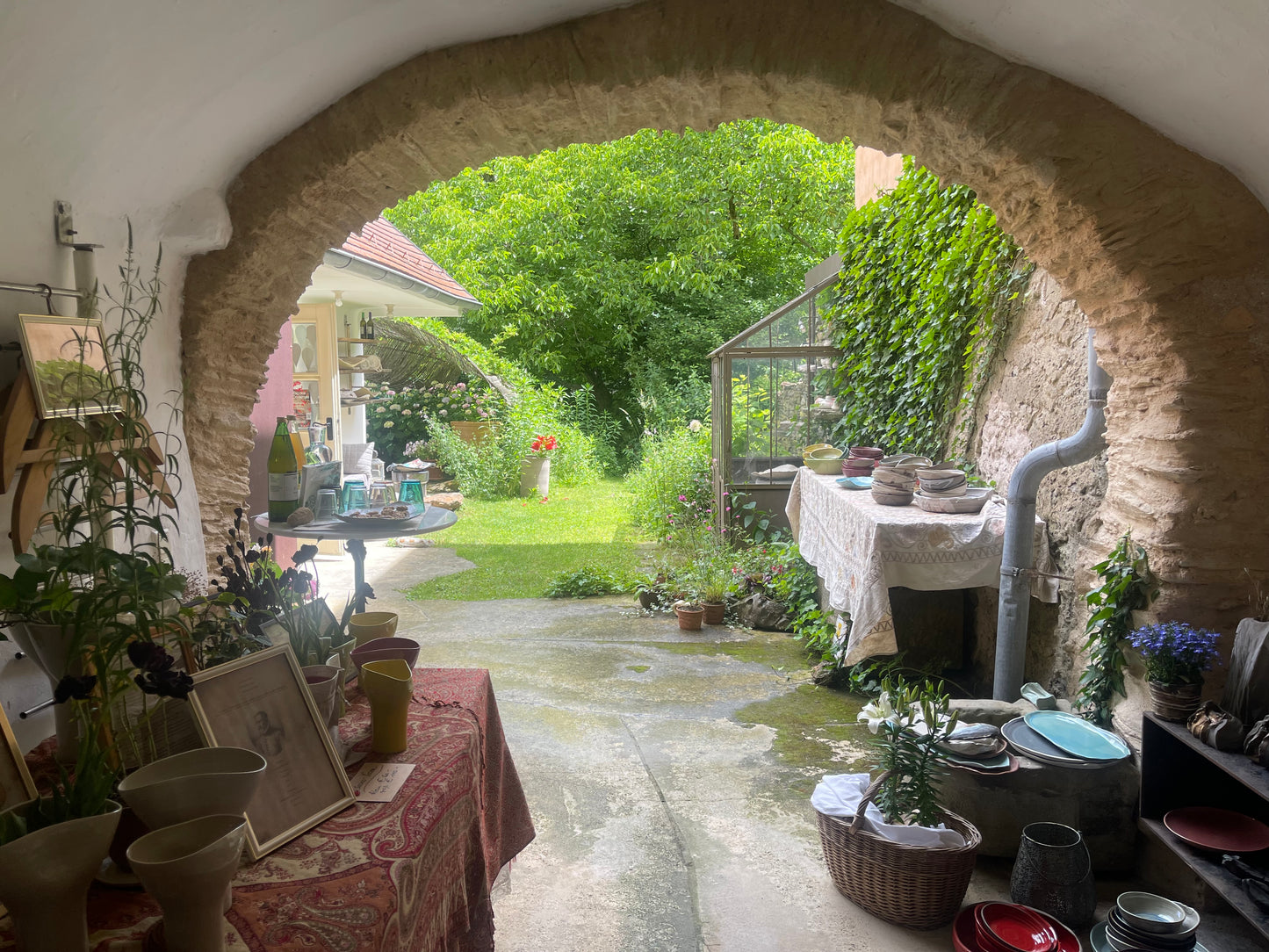 A rustic stone archway frames an outdoor garden patio with green grass, trees, and plants. Tables on both sides display pottery, vases, framed art, and kitchenware, creating a cozy, home-style display.
