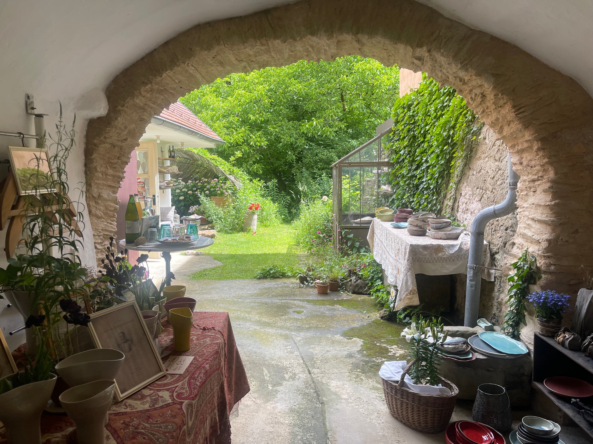 A rustic stone archway frames an outdoor garden patio with green grass, trees, and plants. Tables on both sides display pottery, vases, framed art, and kitchenware, creating a cozy, home-style display.
