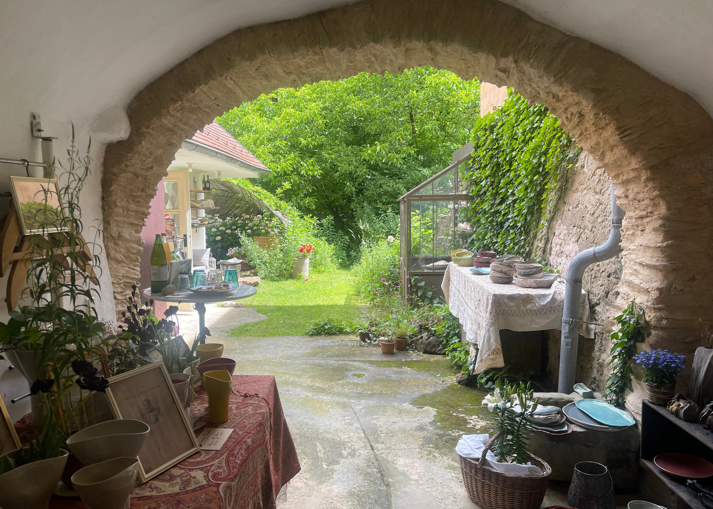 A rustic stone archway frames an outdoor garden patio with green grass, trees, and plants. Tables on both sides display pottery, vases, framed art, and kitchenware, creating a cozy, home-style display.