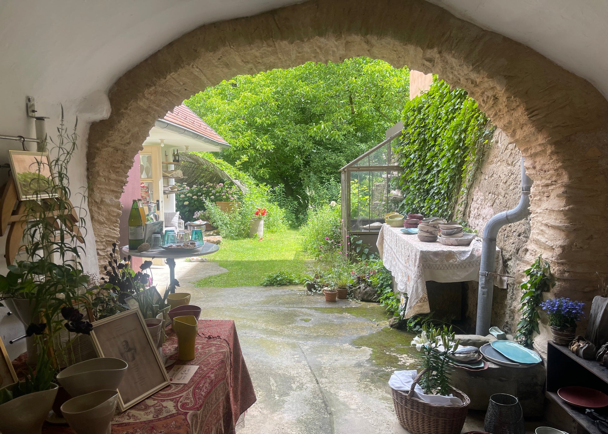 A rustic stone archway frames an outdoor garden patio with green grass, trees, and plants. Tables on both sides display pottery, vases, framed art, and kitchenware, creating a cozy, home-style display.