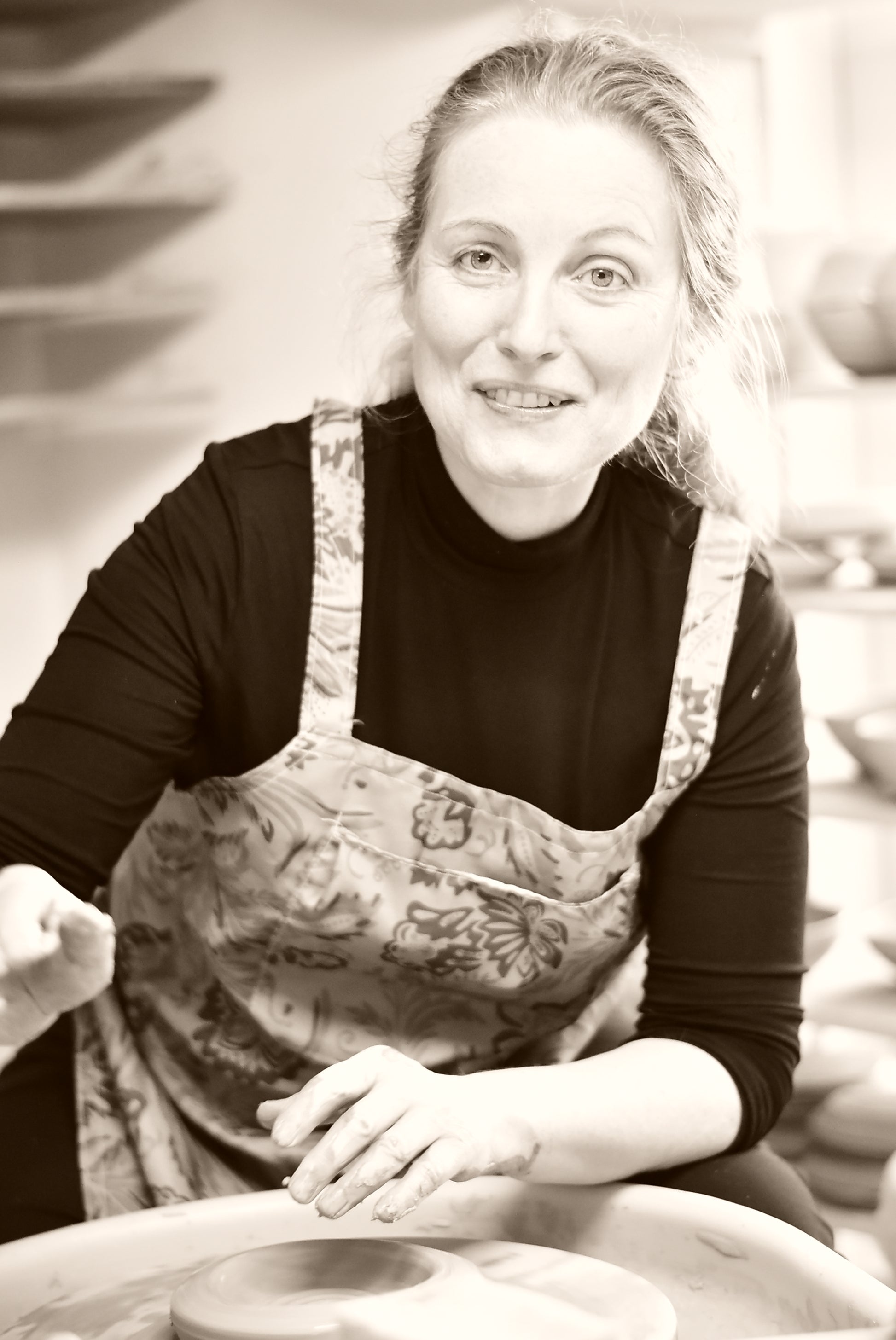 A woman with blonde hair tied back, wearing a black long-sleeve shirt and a floral apron, smiling while working at a pottery wheel in a studio.