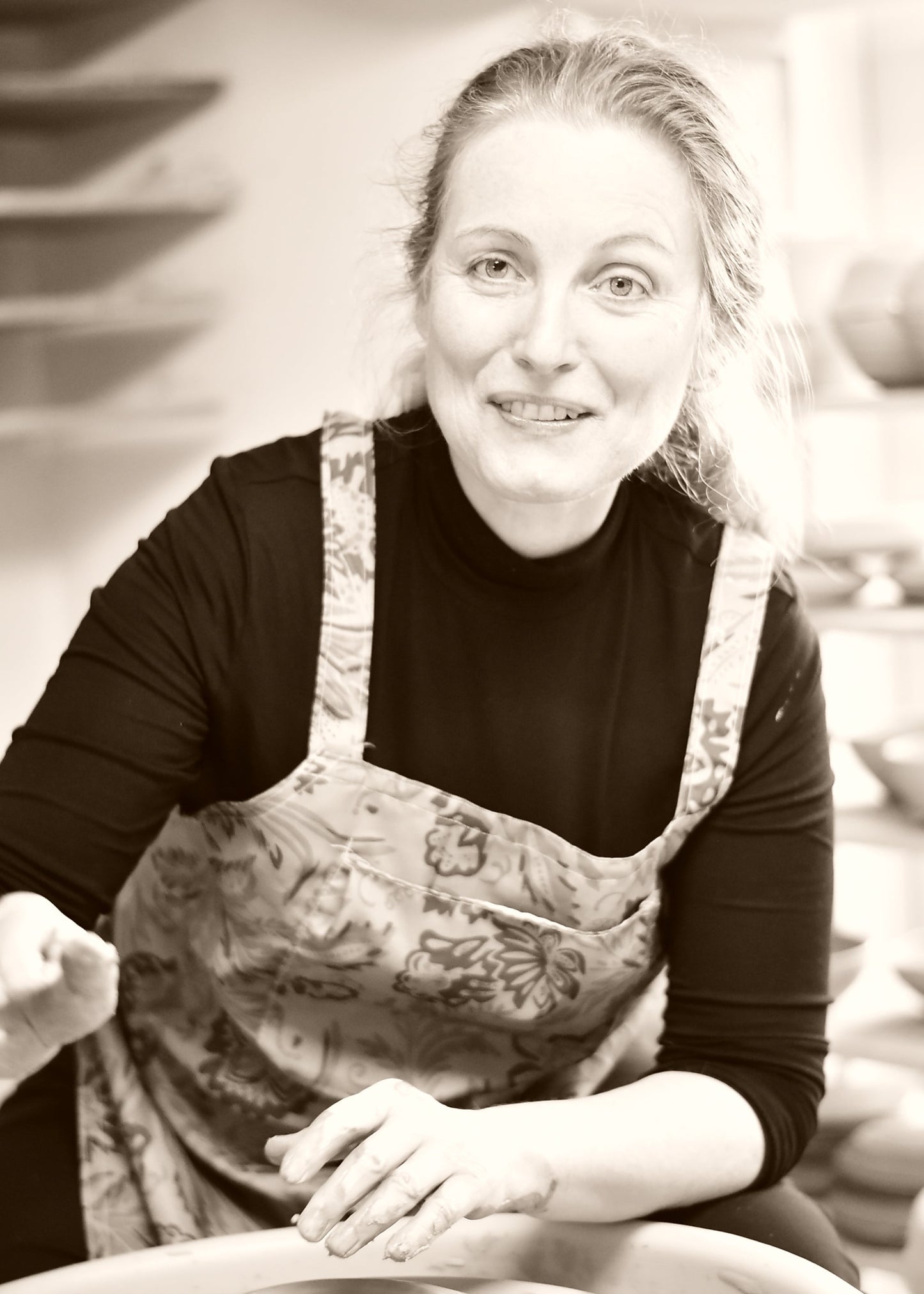 A woman with blonde hair tied back, wearing a black long-sleeve shirt and a floral apron, smiling while working at a pottery wheel in a studio.