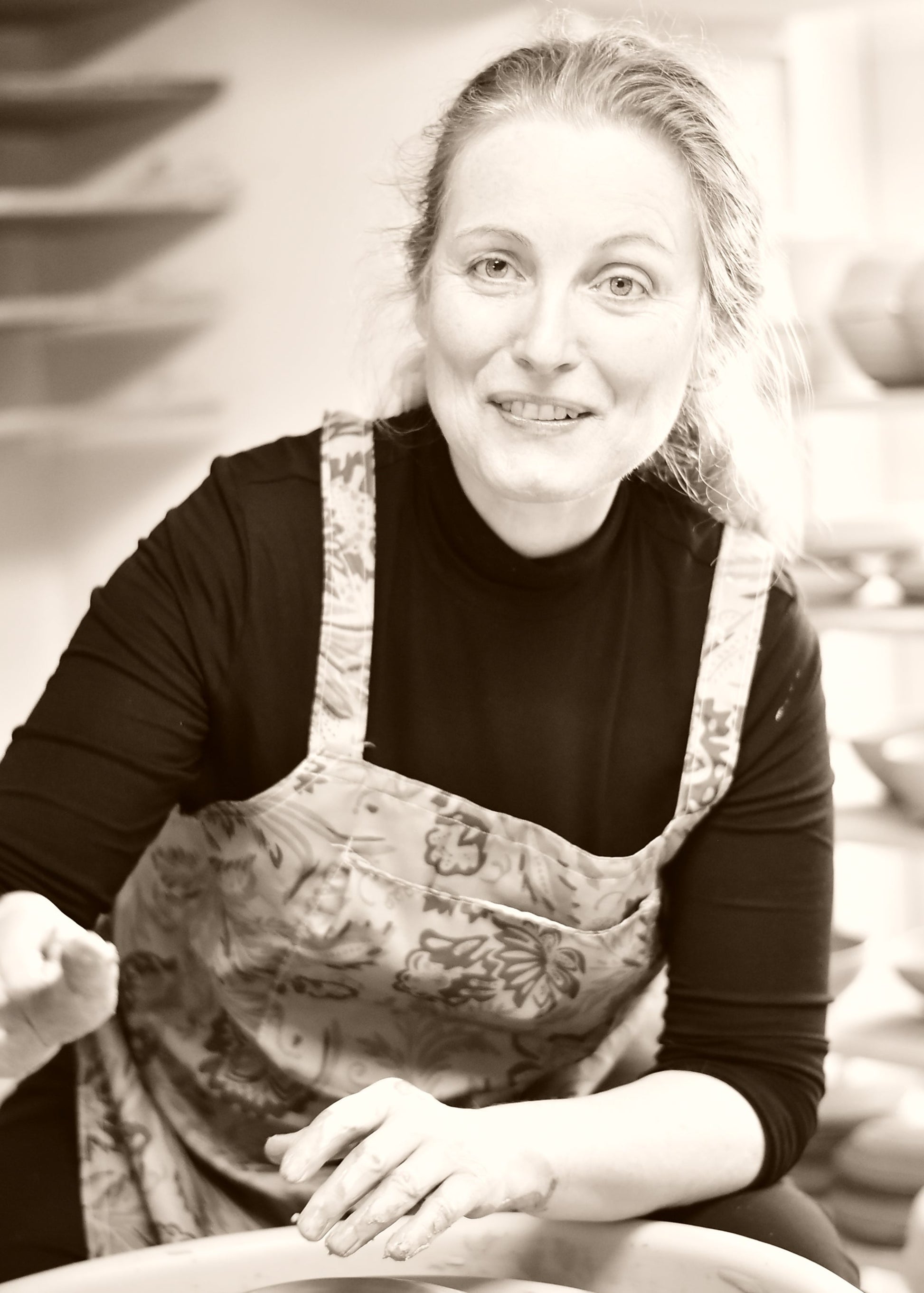 A woman with blonde hair tied back, wearing a black long-sleeve shirt and a floral apron, smiling while working at a pottery wheel in a studio.