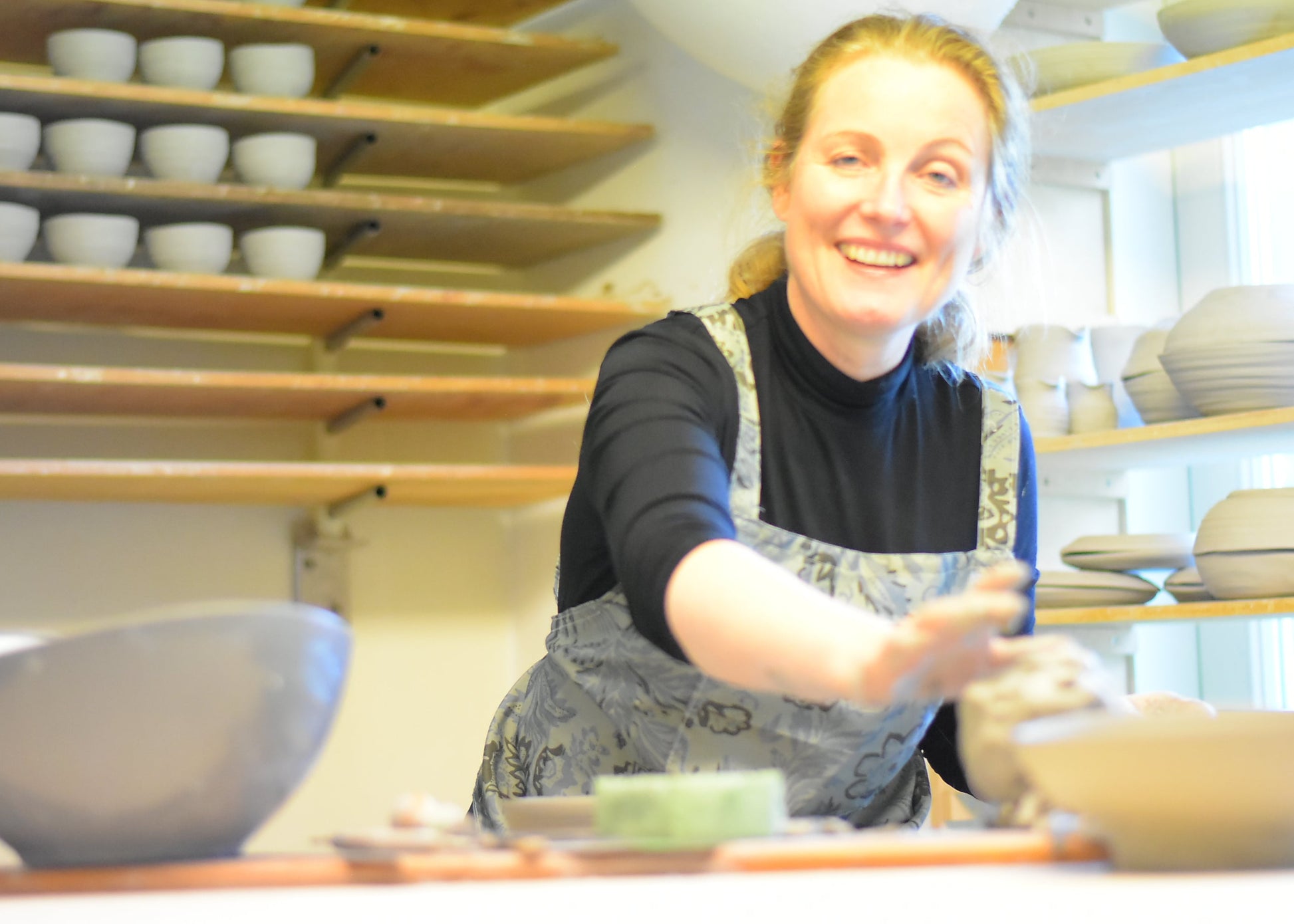 A smiling woman in a pottery studio wearing a black turtleneck and gray patterned apron, reaching toward clay on a table, with shelves of ceramic bowls in the background.