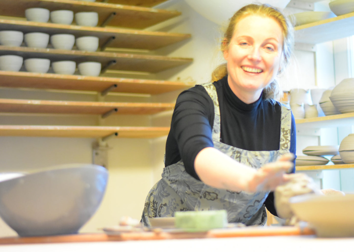 A smiling woman in a pottery studio wearing a black turtleneck and gray patterned apron, reaching toward clay on a table, with shelves of ceramic bowls in the background.