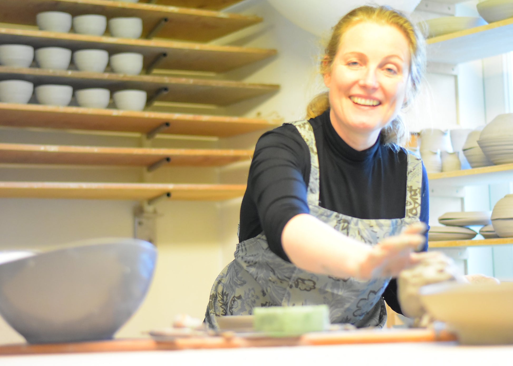 A smiling woman in a pottery studio wearing a black turtleneck and gray patterned apron, reaching toward clay on a table, with shelves of ceramic bowls in the background.