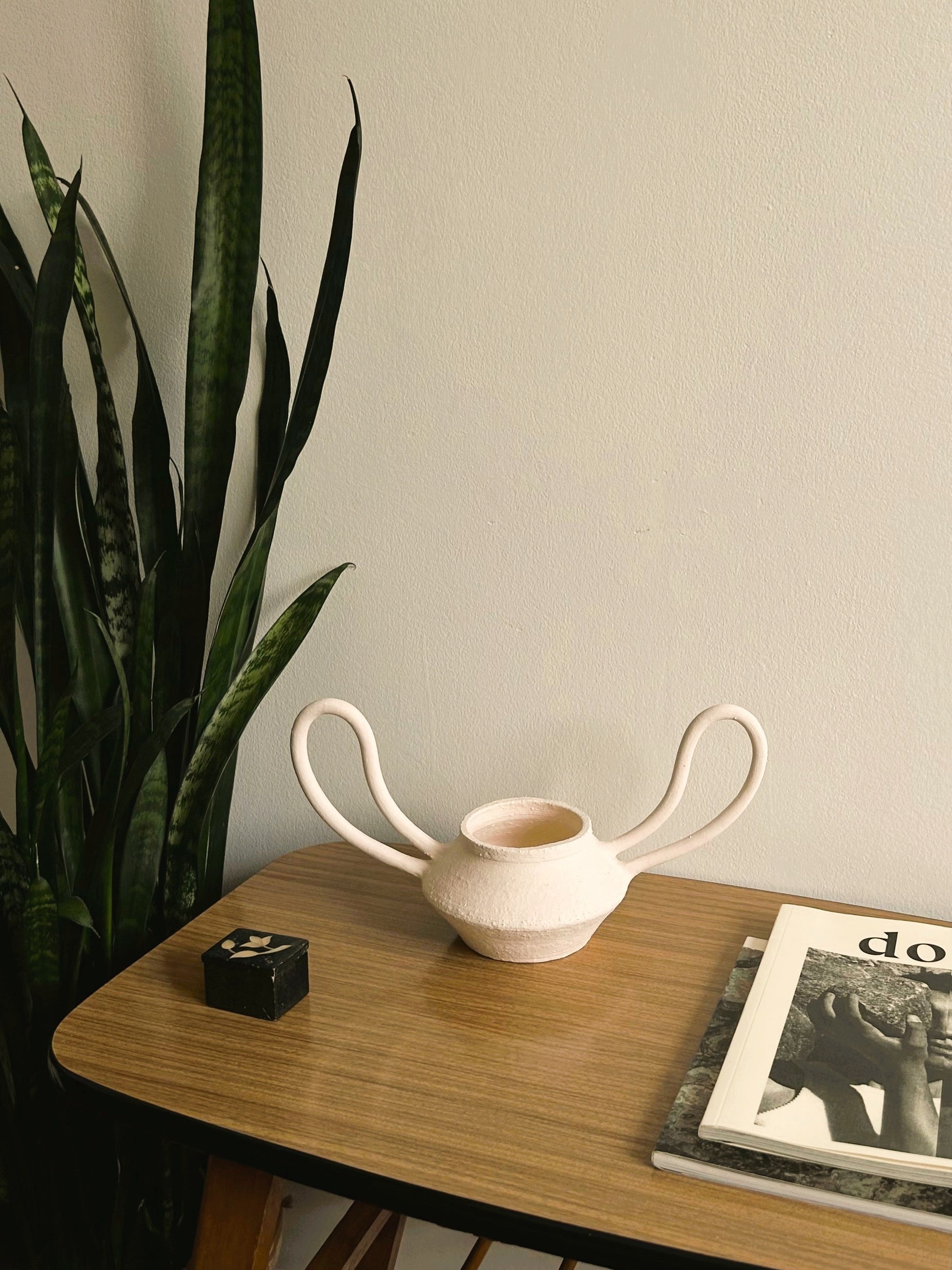 White ceramic vase on a wooden surface with a plant and magazine in the background