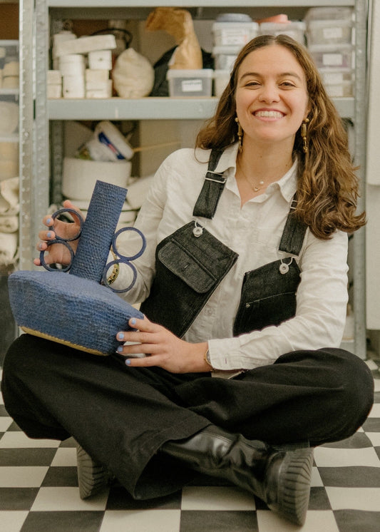 Person sitting on a checkered floor holding a blue bag and black overalls in a storage room.
