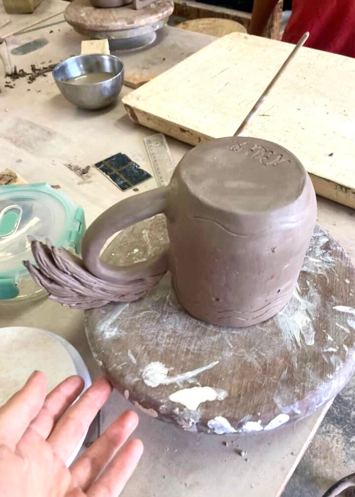 Clay pot on a pottery wheel with a hand guiding it, surrounded by pottery tools and materials.