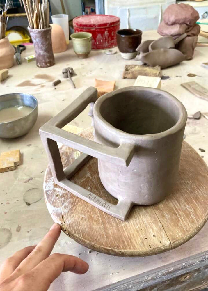 Handmade ceramic mug on a pottery wheel with a hand pointing at it, surrounded by pottery tools and materials.