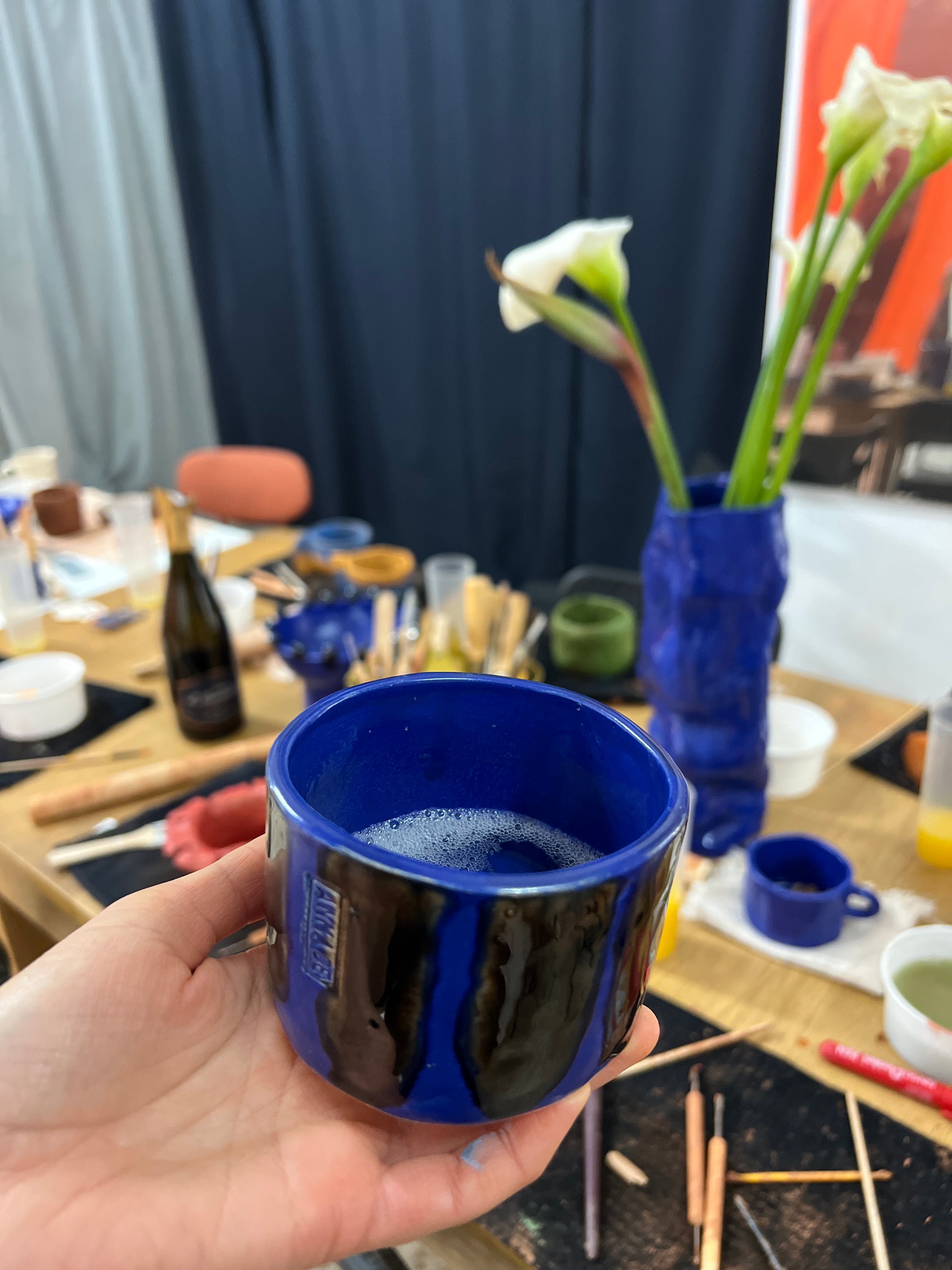 Hand holding a blue cup with a blurred background of a person and various items.