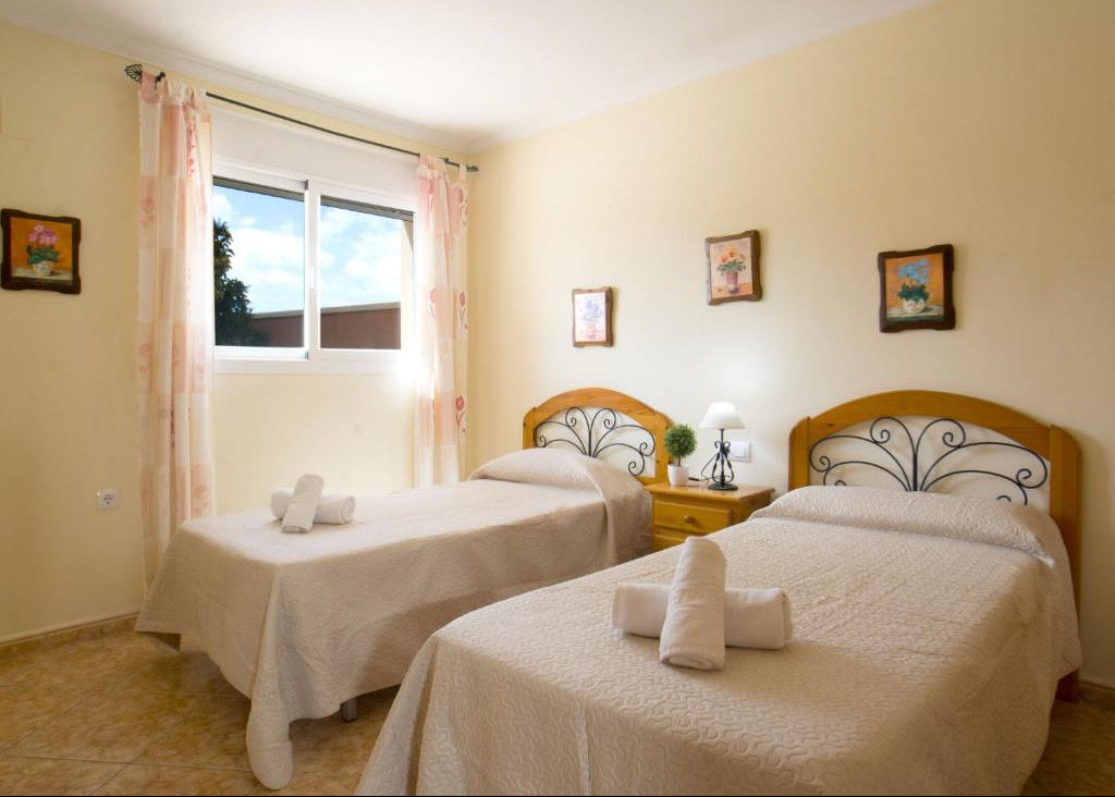 A twin bedroom with two single beds dressed in cream-colored bedding, featuring wooden and wrought-iron headboards. The room has pale yellow walls, a small wooden nightstand between the beds, and a window with pink patterned curtains on the left.