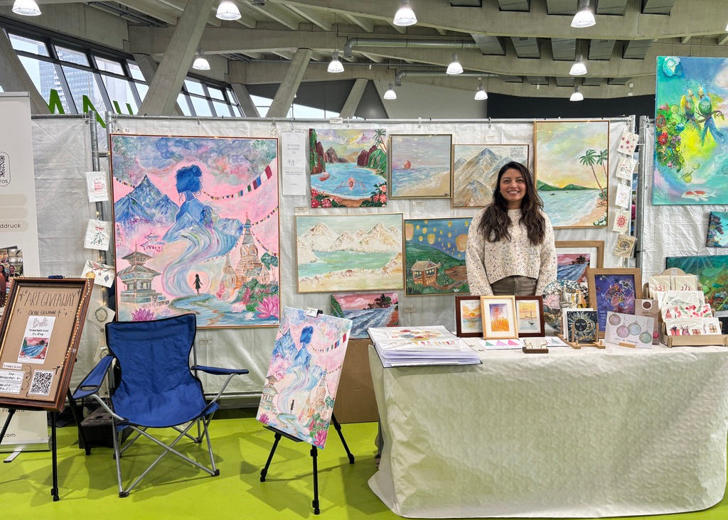 A woman stands behind a table at an art fair booth, surrounded by colorful paintings, framed prints, and art supplies. The booth has a white table covered with a light cloth, a blue folding chair, and several art pieces displayed on easels and walls in a large indoor exhibition space.