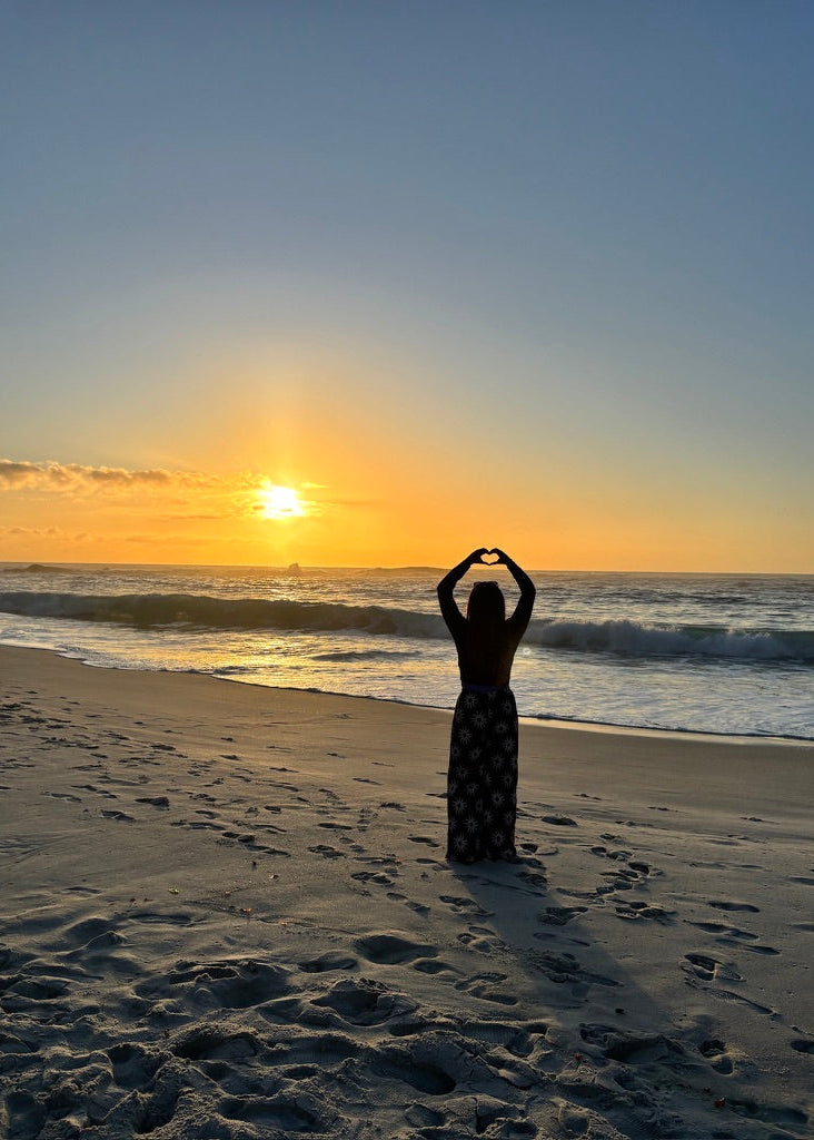 A woman stands on a sandy beach at sunset facing the ocean, forming a heart shape with her hands. She is wearing a long dark dress with a light pattern.