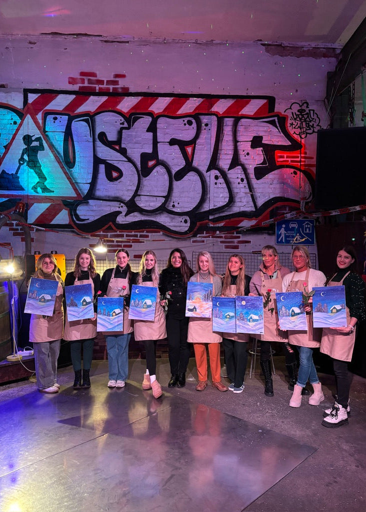 A group of ten people standing together in a studio or workshop, each holding a blue and white book or portfolio. They are positioned in front of a graffiti-covered brick wall with the word 'BUSTLE' prominently displayed.