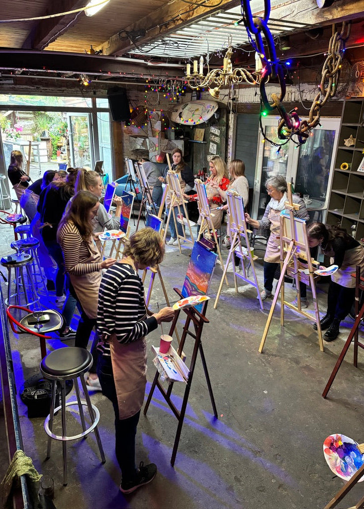A group of people painting at easels inside a creative studio or art workshop with shelves, hanging decor, and a rustic industrial ceiling.