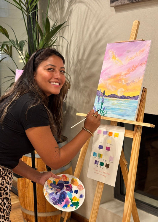 A woman painting a colorful landscape scene on a canvas set on an easel, holding a paint palette with mixed colors. She is smiling and looking at the camera in an indoor setting with a plant and wall shadows behind her.
