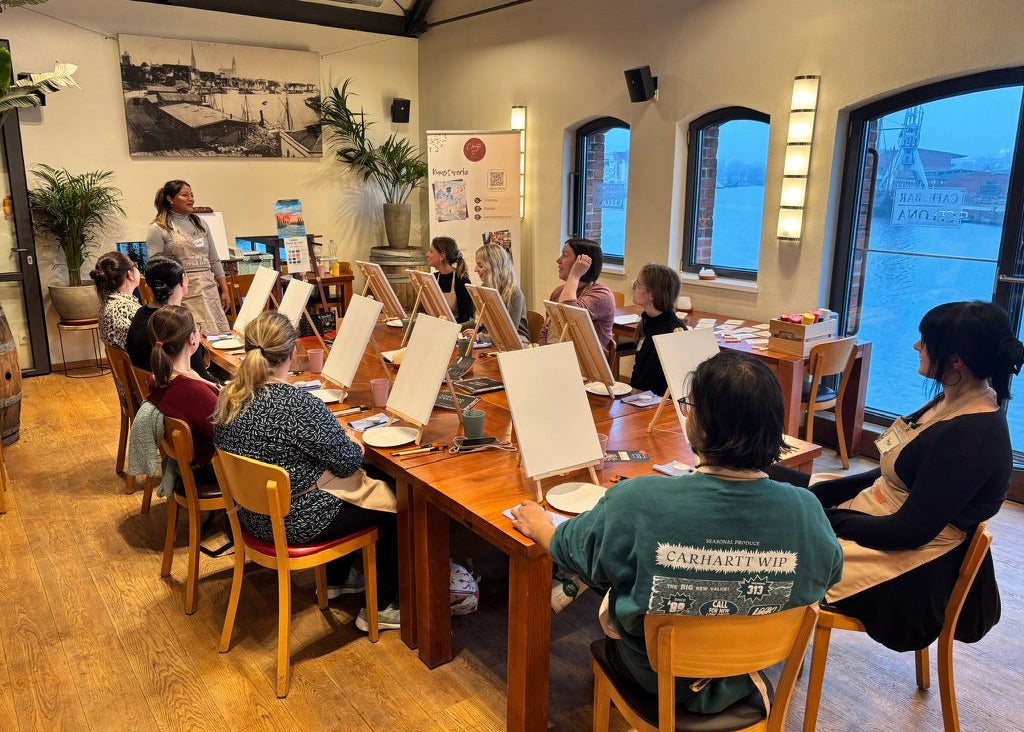 A group of people seated at long wooden tables in a bright indoor studio or classroom, facing a presenter standing near the front. Each person has an easel or tabletop setup in front of them, with large windows, plants, and framed wall art visible in the room.