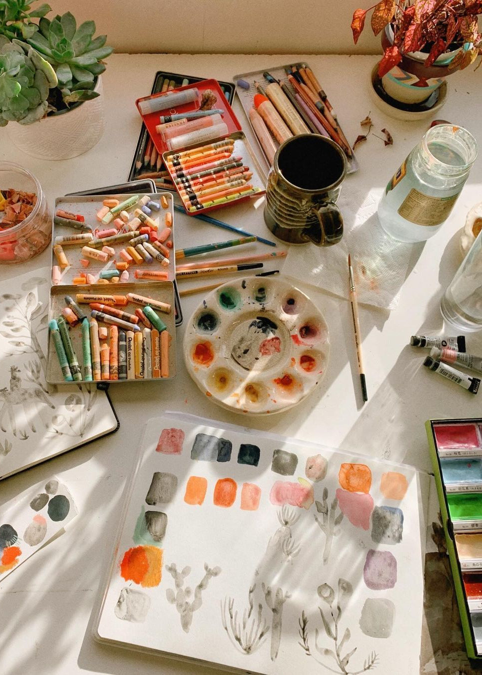 A styled flat-lay of an artist's studio setup on a sunlit white table, featuring multiple boxes of colored pencils, watercolor paints, paint tubes, brushes, a palette with mixed colors, sketchbooks, and small plants.