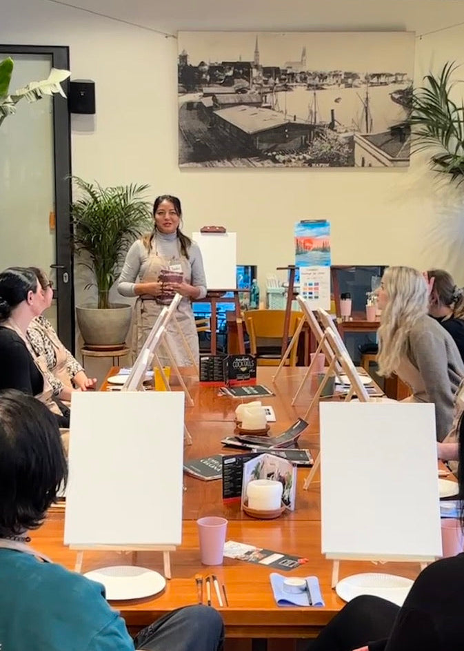 A group of people seated around a wooden table in a bright indoor studio or meeting space, facing a woman standing behind a whiteboard. Several blank white presentation boards on easels are arranged on the table, along with cups, papers, and a large black-and-white cityscape print on the wall behind.