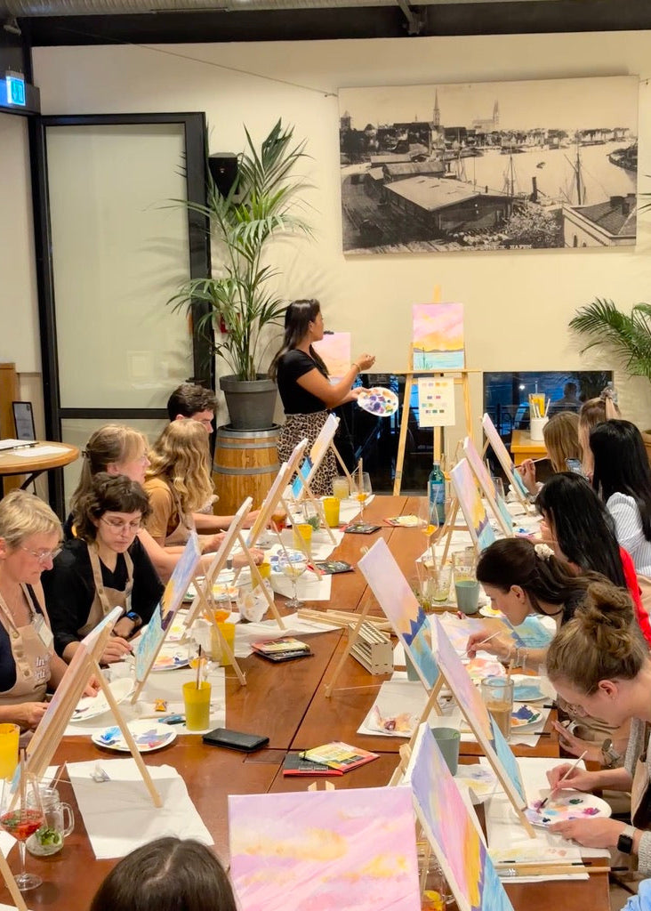 A group of people seated at a long wooden table, painting on small easels in a bright indoor studio or workshop. A woman stands at the front holding a paint palette, with paintings, bottles, plants, and a large black-and-white harbor scene print on the wall in the background.
