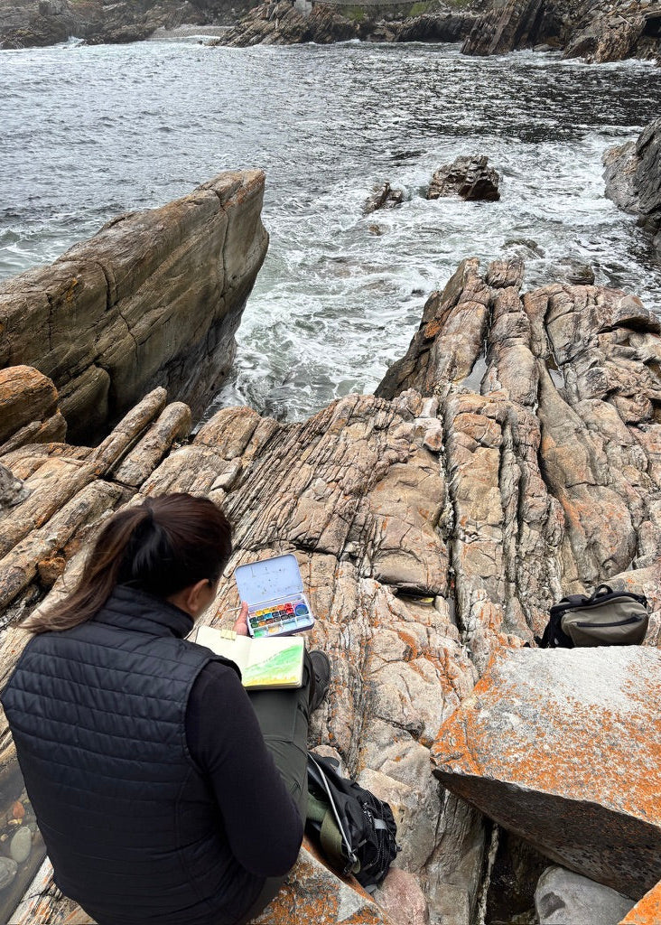A person sitting on rocky coastal ground by the ocean, facing away from the camera, with a notebook and a small colorful item on their lap. The background shows waves crashing against layered tan and gray rocks.