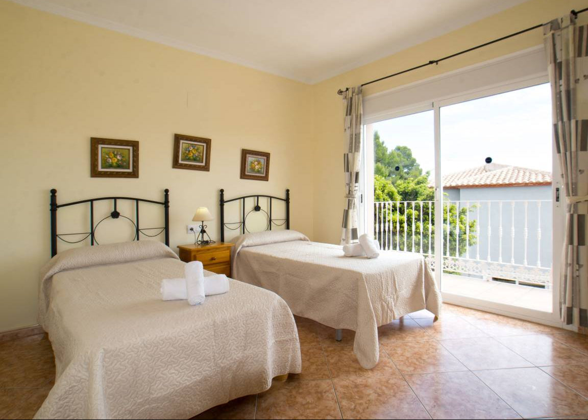A twin bedroom with two single beds dressed in beige linens, black metal headboards, and white towels, set against pale yellow walls with a sliding glass door leading to a balcony.