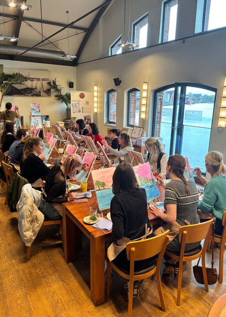 A group of people seated at long wooden tables in a bright indoor studio or cafe, painting on canvases with easels. Large windows and arched glass doors overlook water, and the room has high ceilings with exposed beams.