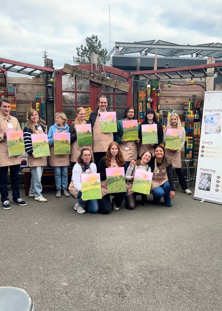 A group of people posing outdoors in front of a workshop or garden center, each holding a small colorful landscape painting. Several people are wearing tan aprons, and a white vertical sign stands to the right.