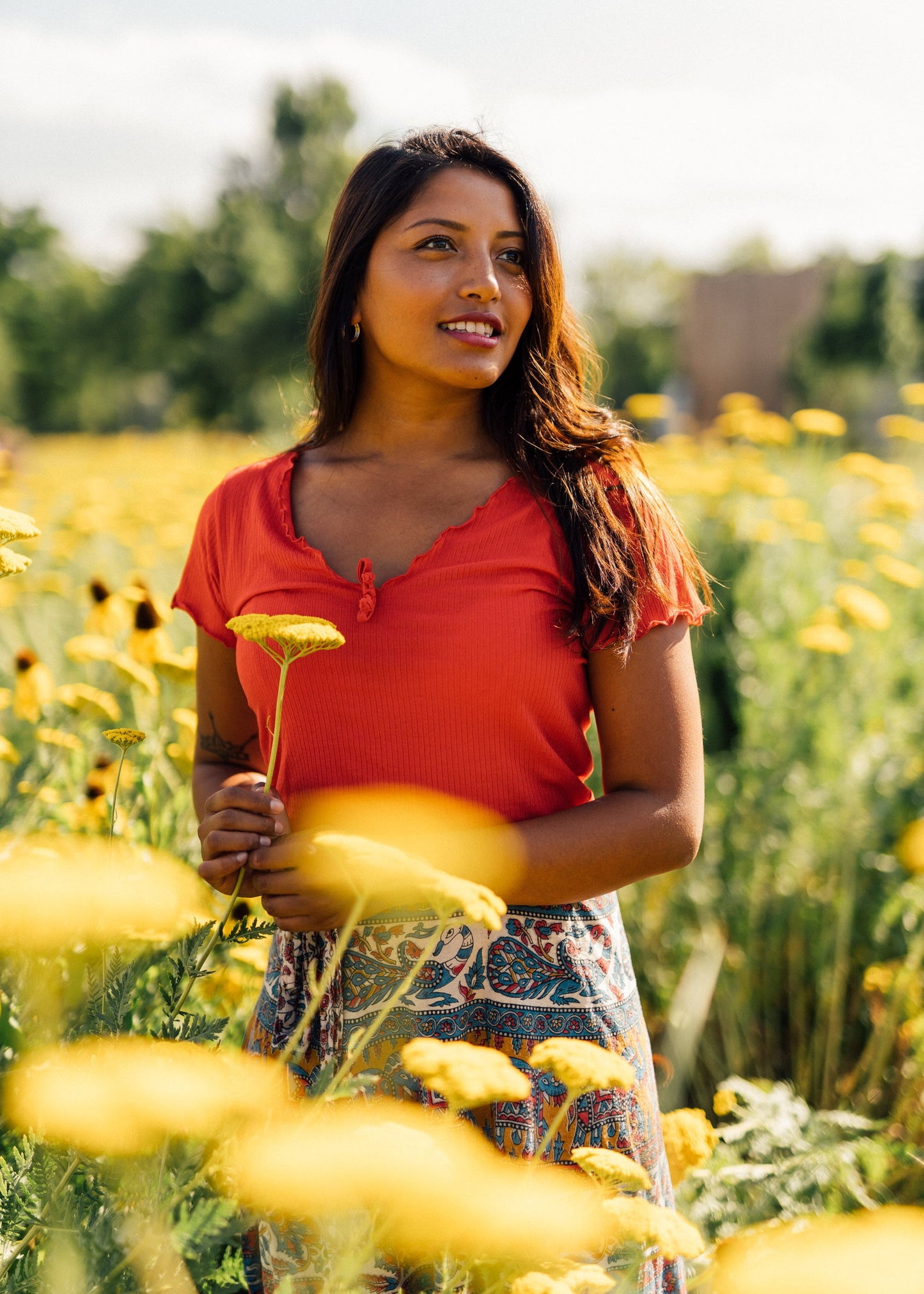 A woman standing in a field of yellow flowers, wearing a red short-sleeve top and a patterned skirt, holding a yellow flower.