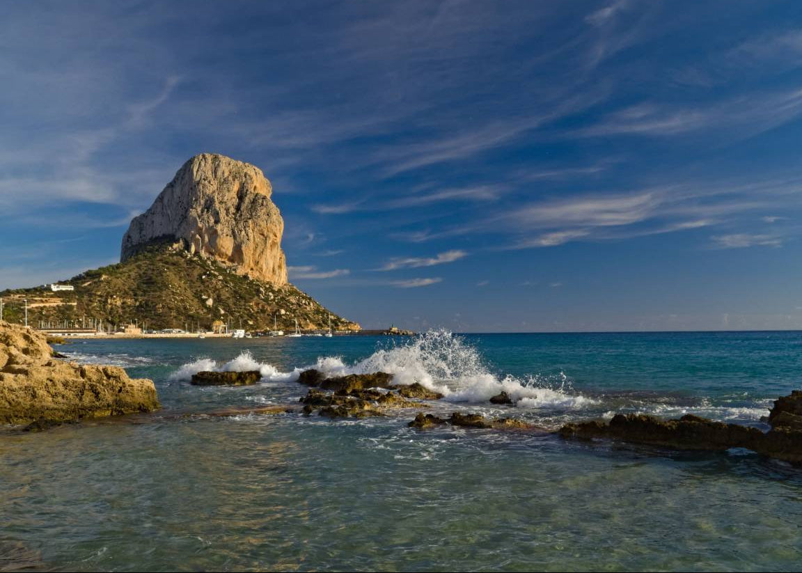 A scenic coastal landscape with a large rock formation rising from the sea under a blue sky with wispy clouds, turquoise water, and white surf breaking around dark rocks in the foreground.