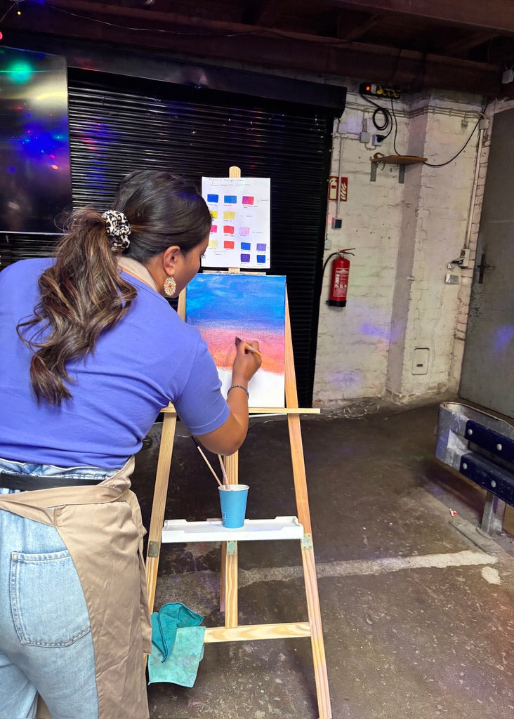 A woman in a purple shirt and beige apron paints on a canvas set up on a wooden easel in an indoor studio space with a black roller shutter and concrete walls.