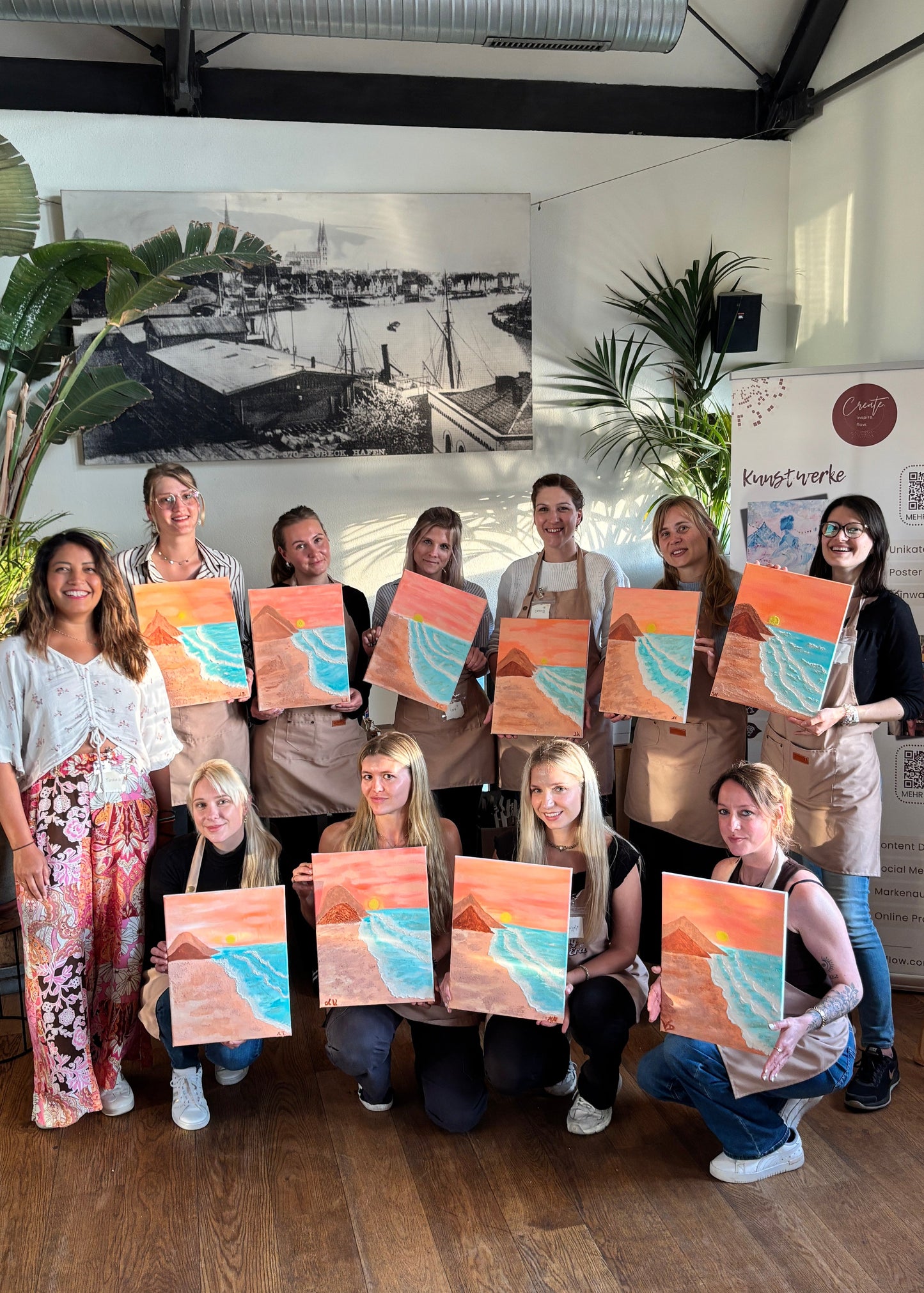 A group of women holding colorful paintings in an indoor studio or workshop space with wood floors, plants, and a large black-and-white wall print.