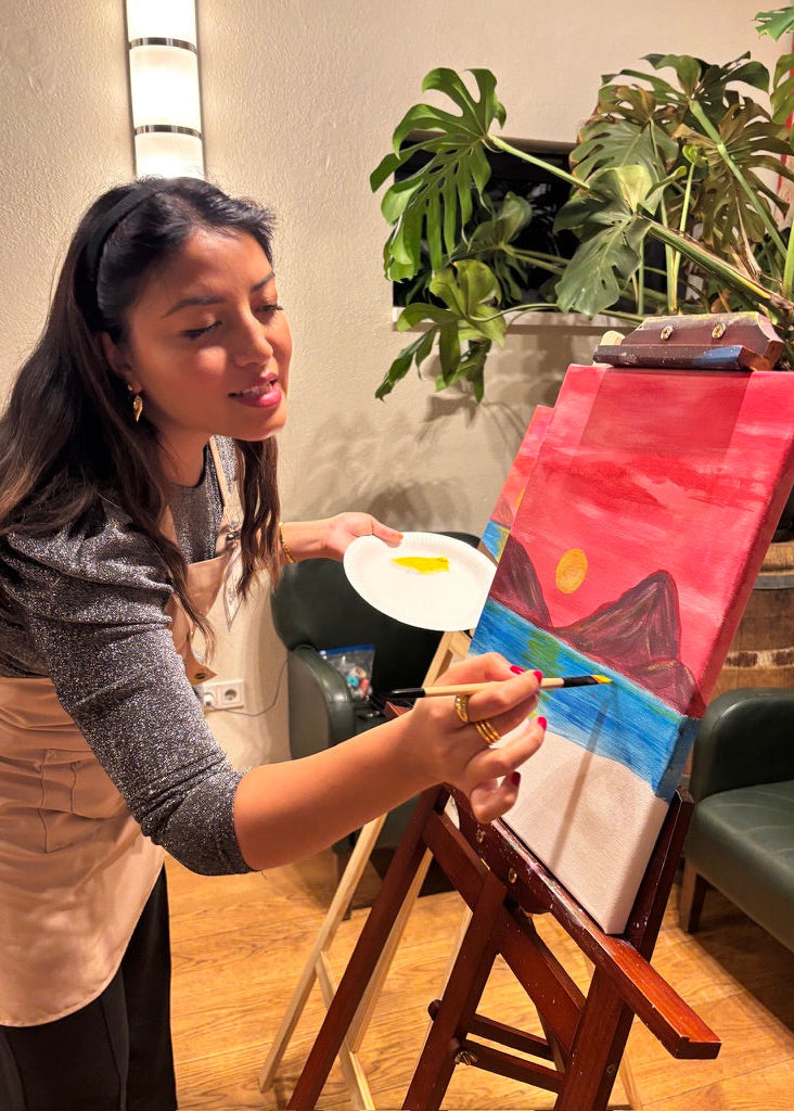 A woman wearing a beige apron over a gray long-sleeve top is painting on a canvas set on an easel. The canvas shows a colorful landscape with red sky, blue water, and dark mountains.