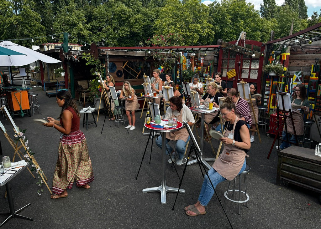 An outdoor art studio or workshop scene with multiple people seated at small tables and easels, painting or drawing in a casual, creative environment. Trees, tables, chairs, and art supplies are visible throughout the space.