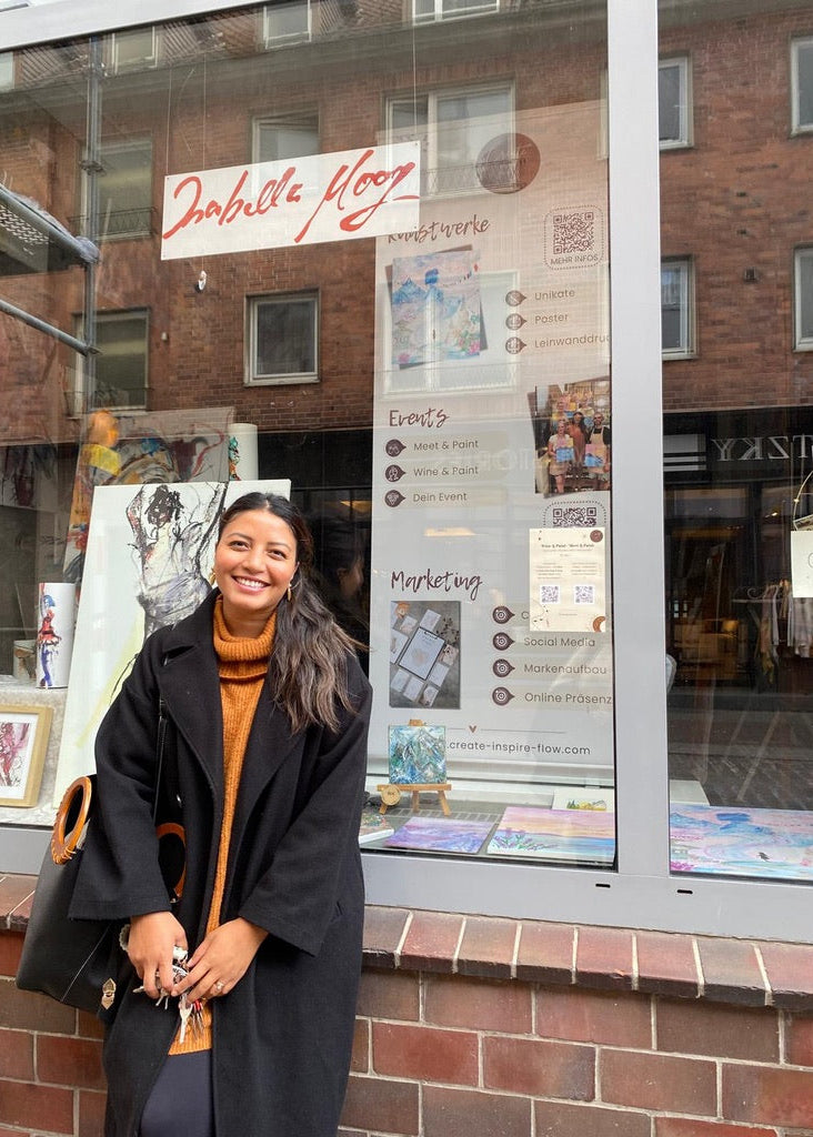 A woman smiling in front of a storefront window with a brick wall. She is wearing a black coat over a rust-orange turtleneck sweater and holding a black shoulder bag.